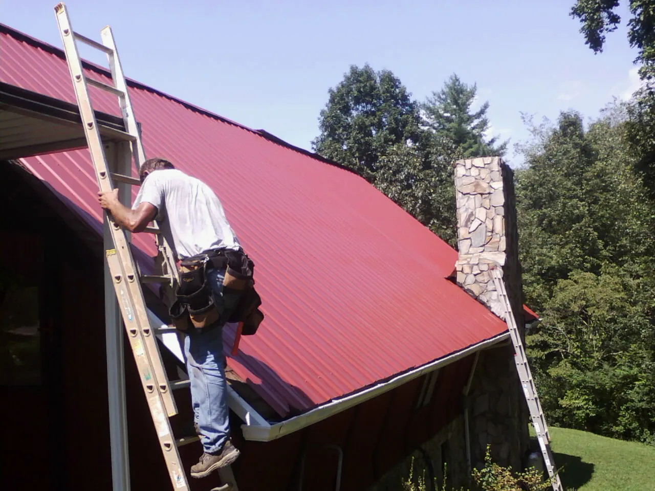 Roofer on a ladder beside a red metal roof and stone chimney — Soffit & Fascia Repair by Roofing Brothers in Cannon Falls